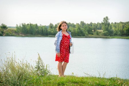 Téléchargez les photos : En été, une fille vêtue d'une veste en jean et d'une robe rouge se tient sur la rive de la rivière. - en image libre de droit
