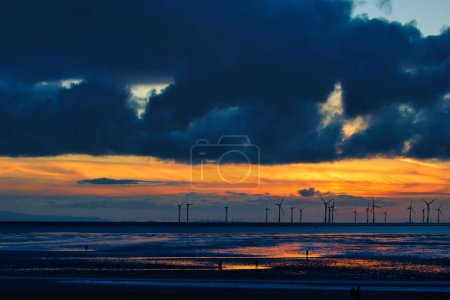 Dramatic sunset over a beach with silhouetted wind turbines on the horizon and reflective wet sand in Crosby, England.