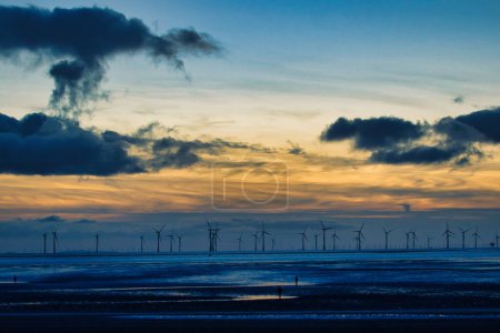 Heiterer Sonnenuntergang über einem Windpark mit Silhouetten von Turbinen und dramatischen Wolken, die sich auf dem Wasser in Crosby, England, spiegeln.