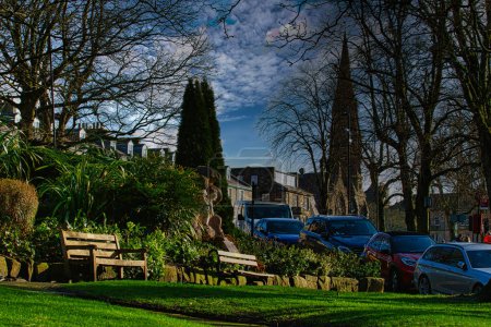 Tranquila escena del parque urbano con bancos y exuberante vegetación, ambientada sobre un telón de fondo de edificios históricos y cielo azul con nubes en Harrogate, Inglaterra.