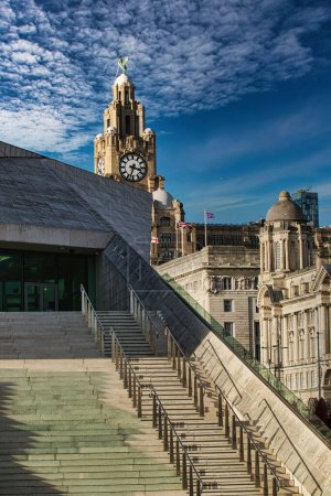 Escalera moderna que conduce a la histórica torre del reloj bajo un cielo azul con nubes tenues en Liverpool, Reino Unido.