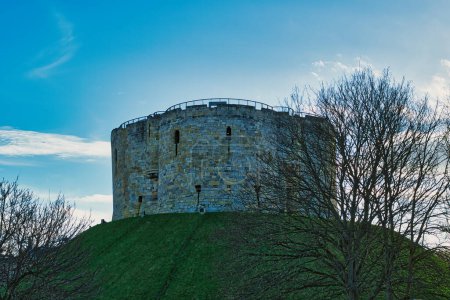 Tour médiévale en pierre sur une colline herbeuse avec des arbres nus contre un ciel bleu avec des nuages à York, Royaume-Uni.
