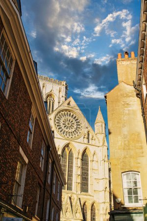 Historic cathedral facade with rose window, framed by old buildings against a blue sky with clouds in York, UK.