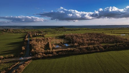 Aerial view of lush green fields with a small forest area, under a vast blue sky with fluffy clouds in North Yorkshire.