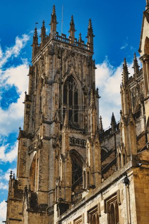 Gothic cathedral tower against a blue sky with clouds, showcasing intricate architectural details and flying buttresses in York, North Yorkshire, England.