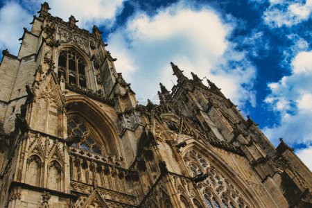 Angle dramatique de la façade d'une cathédrale gothique avec des sculptures complexes en pierre sur un ciel bleu vif avec des nuages duveteux, mettant en valeur la grandeur architecturale et l'élégance historique à York, dans le Yorkshire du Nord, en Angleterre.
