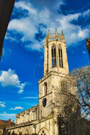 Gothic church tower against a vibrant blue sky with fluffy clouds, showcasing intricate architectural details and a blooming tree at the corner in York, North Yorkshire, England.