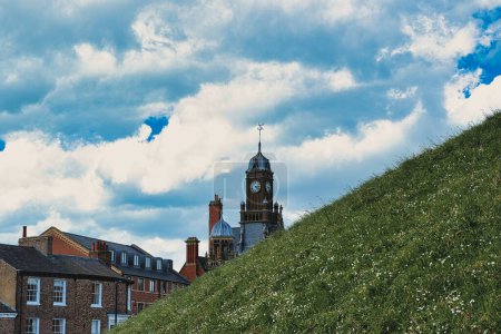 Quaint European town with historic buildings and a clock tower, set against a vibrant blue sky with fluffy clouds, and a lush green hill in the foreground in York, North Yorkshire, England.