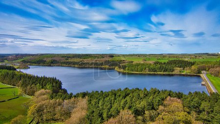 Vista aérea de un lago sereno rodeado de exuberantes bosques verdes bajo un cielo azul vibrante con nubes dispersas.