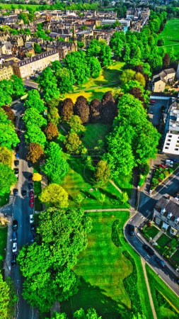 Aerial view of a lush green park surrounded by residential buildings and roads. The park features various trees and open grassy areas, with a few people visible enjoying the space. The surrounding buildings are a mix of historical and modern architec