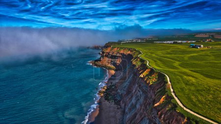 Aerial view of a dramatic coastal cliff with green fields on top, a winding path along the edge, and a misty ocean below. The sky is filled with swirling clouds, creating a dynamic and picturesque scene.