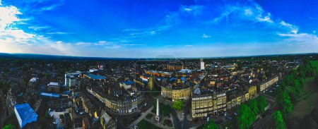 Luftaufnahme einer Stadt mit einer Mischung aus historischen und modernen Gebäuden, umgeben von viel Grün unter einem klaren blauen Himmel.