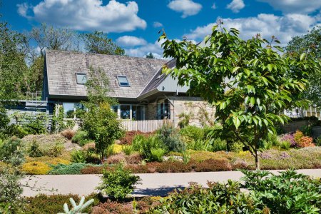 Une maison moderne avec un toit en pente entouré d'un jardin luxuriant et paysager avec diverses plantes et arbres. Le ciel est clair avec quelques nuages.