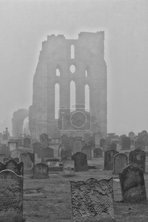 A foggy scene of an old, weathered cemetery with numerous gravestones in the foreground. In the background, the ruins of a large, ancient stone building are visible, partially obscured by the mist.