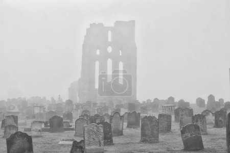 A foggy cemetery with old tombstones and the ruins of a large building in the background.