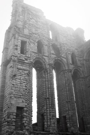 Eine Schwarz-Weiß-Fotografie einer antiken Steinruine mit hohen Bogenfenstern und verwittertem Mauerwerk. Das Bauwerk scheint Teil eines historischen Gebäudes oder einer Burg zu sein.