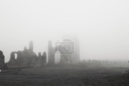 A foggy landscape featuring the ruins of an ancient stone building, with arches and walls partially visible through the dense mist. The scene is eerie and atmospheric, evoking a sense of mystery and history.