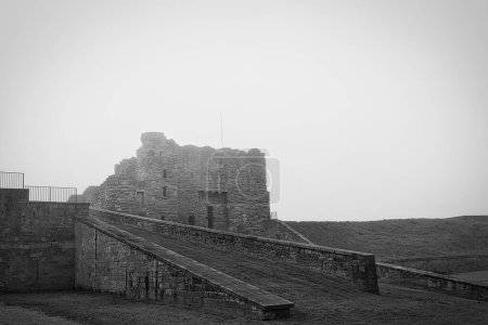 A black and white photograph of an old stone castle with a ramp leading up to it. The sky is overcast, adding a moody atmosphere to the scene.