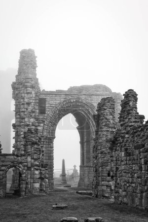 A black and white photograph of ancient stone ruins with an archway, surrounded by fog. The ruins are weathered and partially collapsed, with a grassy area in the foreground.