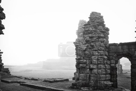 Black and white photograph of ancient stone ruins shrouded in fog. The ruins consist of tall, weathered stone structures and arches, with a misty background obscuring further details.
