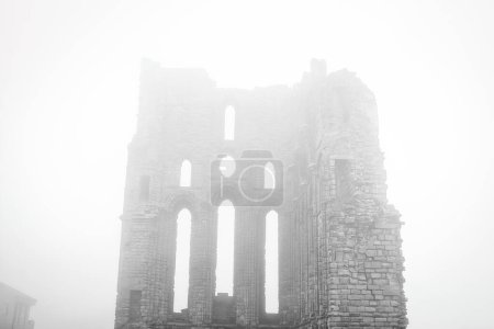 A foggy scene featuring the ruins of an ancient stone building with tall, narrow windows and arches. The structure appears to be partially collapsed, and the dense fog obscures the background, giving the image a mysterious and eerie atmosphere.