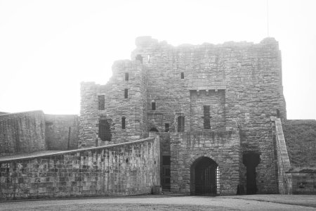 Eine Schwarz-Weiß-Fotografie einer alten steinernen Burg mit einer teilweise zerstörten Struktur. Die Burg hat mehrere Ebenen, gewölbte Tore und eine Steinmauer, die sich von ihr erstreckt. Der Himmel ist bedeckt und sorgt für eine stimmungsvolle Atmosphäre.