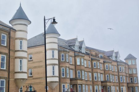 A row of modern brick buildings with unique cylindrical towers and conical roofs. The buildings have multiple windows and are situated under a cloudy sky with a street lamp and a bird in flight.