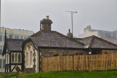 A quaint stone cottage with a fenced yard, surrounded by modern buildings on a foggy day.