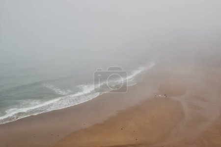A foggy beach scene with waves gently crashing onto the shore. Three people are walking along the wet sand, creating a serene and misty atmosphere.