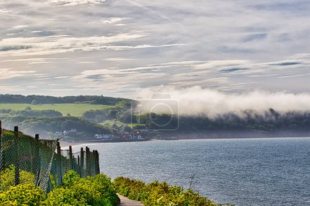 A scenic coastal view with a fence in the foreground, lush green hills, and a small village by the water. The sky is partly cloudy with a layer of mist over the hills.