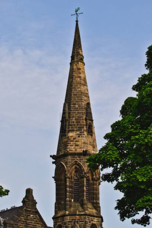 Ein hoher, spitzer Kirchturm aus Stein mit einer Wetterfahne an der Spitze, der vor einem blauen Himmel mit einigen Wolken steht. Grüne Baumblätter rahmen das Bild teilweise ein.