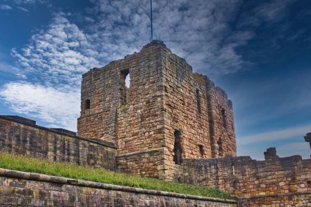 Eine alte steinerne Burgruine mit einem teilweise eingestürzten Turm, vor einem blauen Himmel mit verstreuten Wolken. Die Struktur ist von einer Steinmauer und einer Rasenfläche umgeben.