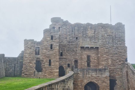 A historic stone castle with weathered walls and a tower, surrounded by a grassy area. The sky is overcast, giving a misty atmosphere.