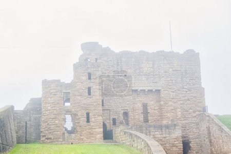Ein nebliger Blick auf eine alte steinerne Burgruine mit teilweise eingestürzten Mauern und einem grasbewachsenen Vordergrund.