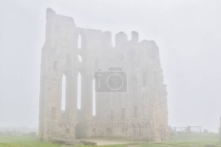 A foggy scene of an ancient stone castle ruin with tall, narrow windows. The structure is partially obscured by the dense fog, creating a mysterious and eerie atmosphere.