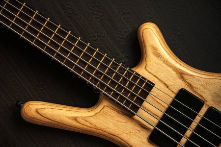 Photo for Close up of electric bass guitar with five strings. Detailed view of wooden varnished texture of musical instrument on dark brown ribbed wooden background. Music classes, concert, hobby and bass player concept background. - Royalty Free Image