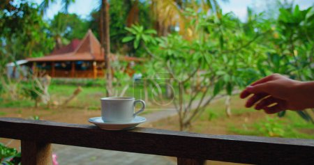 Téléchargez les photos : Jeune femme main prend une tasse de café chaud sur le balcon avec de beaux paysages naturels flous et indonésiennes maisons balinaises traditionnelles sur le fond, au ralenti. - en image libre de droit