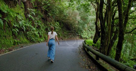 Téléchargez les photos : Woman walks along an asphalt road in a dense forest. Overgrown trees in laurel tree forest or jungle, Anaga National Park, Tenerife, Canary Islands. - en image libre de droit