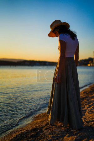 Téléchargez les photos : Athlétique fille caucasienne avec chapeau de paille regardant la mer sur le coucher du soleil sur la plage - en image libre de droit