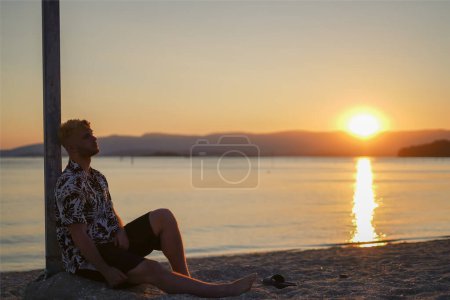 Téléchargez les photos : Jeune homme assis sur le sol regardant le coucher du soleil sur la plage - en image libre de droit