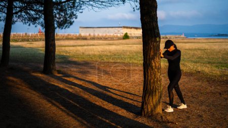 Téléchargez les photos : Jeune fille étirant sa jambe aidant avec un arbre dans le parc - en image libre de droit