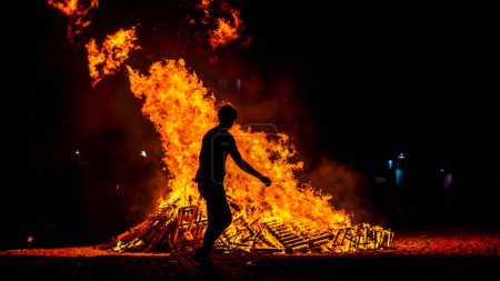 Foto de Hoguera en la playa por la noche con llamas altas con la silueta de un niño bailando en una celebración - Imagen libre de derechos