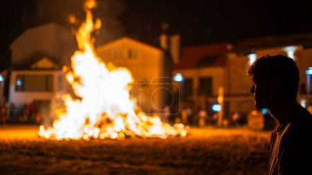 Téléchargez les photos : Célébration rurale avec la ville entière regardant le feu d'un feu de joie et un homme qui le regarde au premier plan à la plage la nuit - en image libre de droit