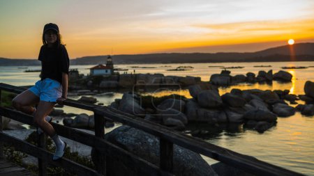 Téléchargez les photos : Jeune fille regardant un chapeau sur le bord de mer debout sur une rampe en bois au coucher du soleil - en image libre de droit