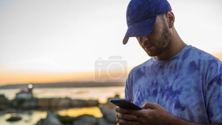 Téléchargez les photos : Jeune homme regardant le smartphone à une belle vue sur la mer au coucher du soleil - en image libre de droit