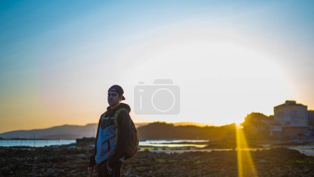 Téléchargez les photos : Jeune homme posant sérieux dans la plage tandis que le coucher du soleil.porter une casquette et un sac à dos - en image libre de droit