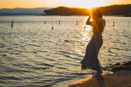 Téléchargez les photos : Jeune belle femme dansant sur la plage bord de mer avec la main contre le coucher du soleil en robe d'été et les vacances - en image libre de droit