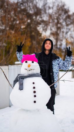 Téléchargez les photos : Joyeux jeune homme avec bonhomme de neige portant un chapeau rose et une écharpe. - en image libre de droit