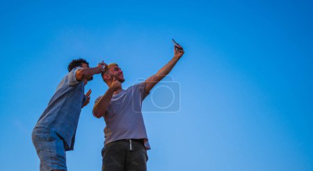 dos amigos multiétnicos tomando una selfie desde una vista de ángulo bajo con el cielo azul en el fondo