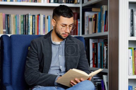 Téléchargez les photos : Homme barbu avec des lunettes assis dans un fauteuil confortable et un livre de lecture, lèvres mobiles, bibliothèques sur fond. Lecteur appréciant son passe-temps à la bibliothèque ou à la librairie. Concept d'éducation - en image libre de droit